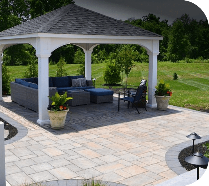 A white gazebo with a dark roof shelters a wicker sectional sofa with blue cushions on a paved patio, surrounded by greenery and potted plants. A black chair and side table sit nearby.