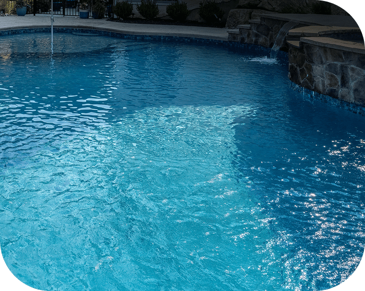 A swimming pool at night with bright blue water, illuminated by underwater lights, creating shimmering reflections on the surface. The pool has a stone-tiled edge and surrounding landscaping.