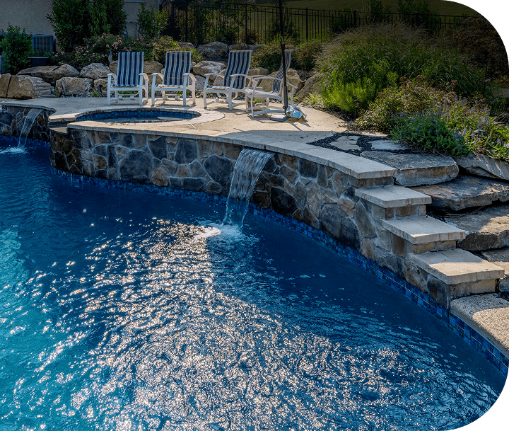 A backyard pool with clear blue water, stone steps, a small waterfall, and a raised hot tub. Four striped lounge chairs are on the pool deck, surrounded by plants and rocks.