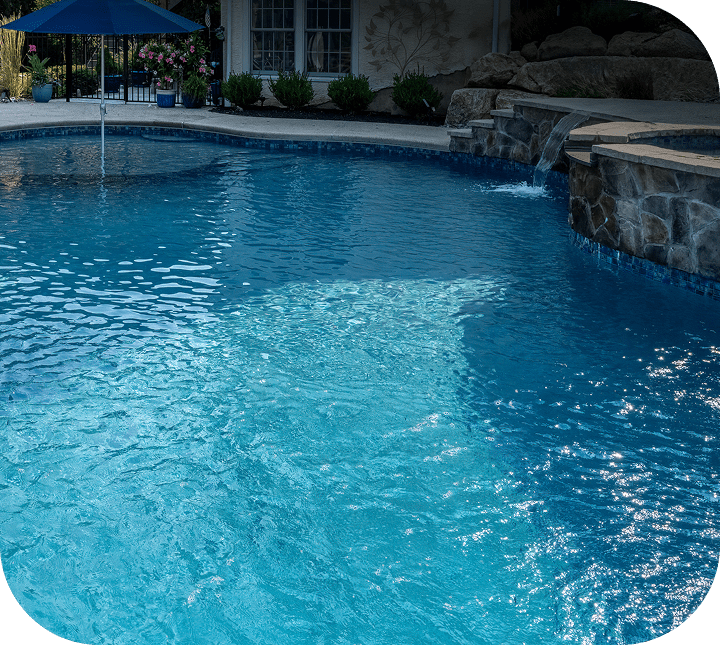 A clear blue swimming pool with a small waterfall feature and stone wall, surrounded by a patio. An umbrella and greenery are visible in the background.