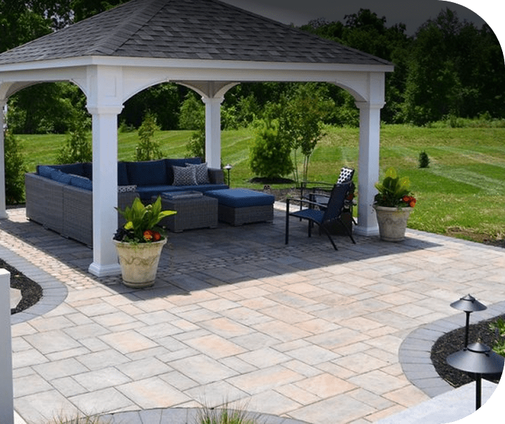 A backyard patio with a white gazebo, featuring gray wicker outdoor furniture with blue cushions, a black chair, two large potted plants, and surrounding green grass and trees.