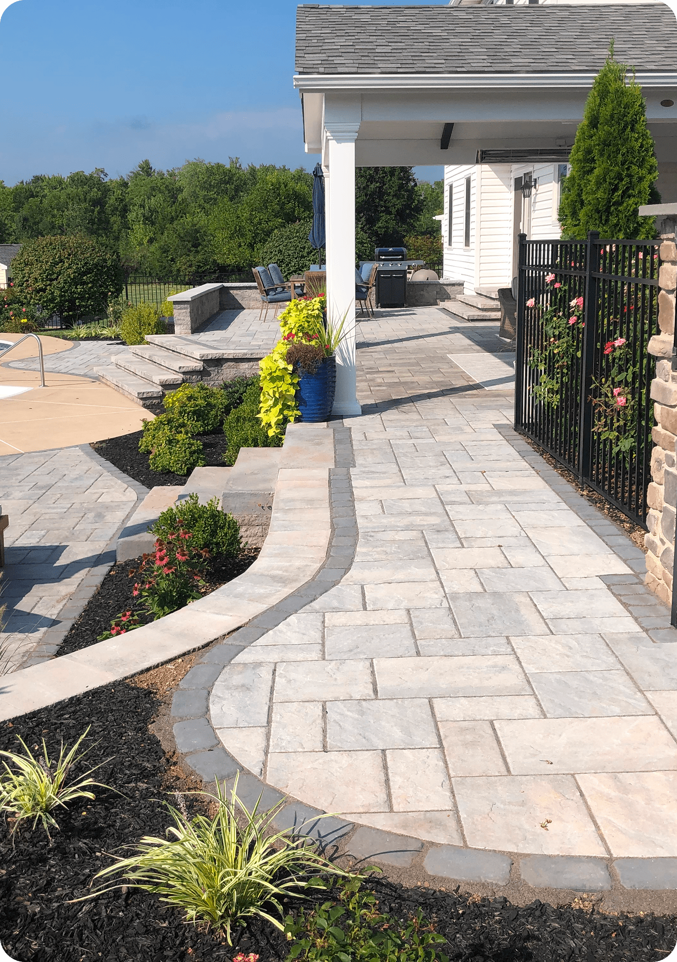 A paved stone walkway curves beside a landscaped garden with shrubs and flowers, leading to a patio area near a white house and a black metal fence. Outdoor seating and a grill are visible under a covered patio.