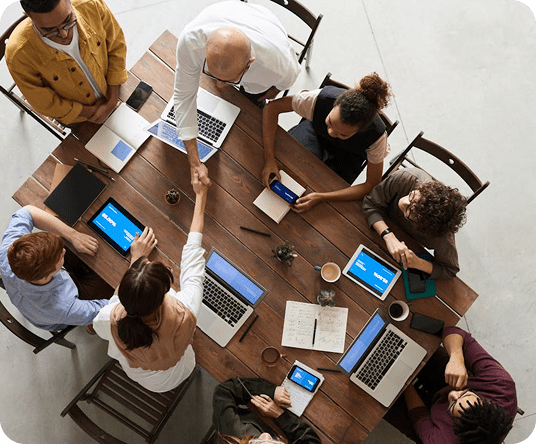 A group of people sit around a wooden table with laptops, notebooks, and coffee cups, as two individuals in the center shake hands, suggesting a collaborative business meeting.