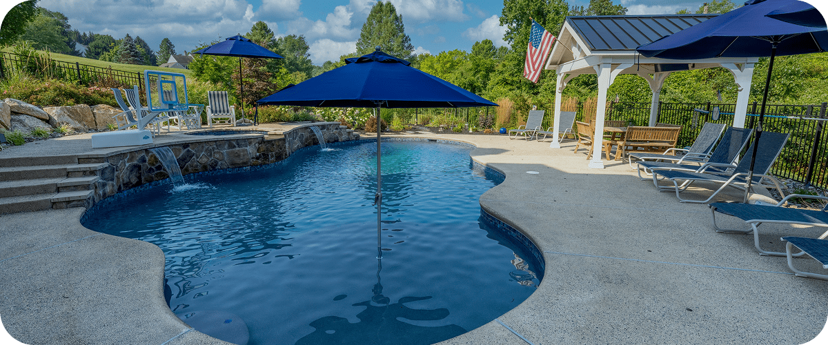 A backyard pool with clear blue water, surrounded by lounge chairs, blue umbrellas, a gazebo, and an American flag, with trees and a grassy hill in the background under a partly cloudy sky.
