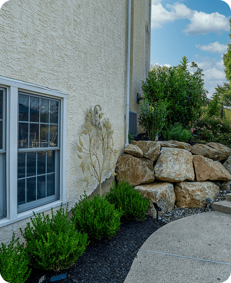 A landscaped area with green shrubs, large stone boulders, and a curved concrete pathway beside a beige stucco house with a window and decorative wall art. Blue sky with clouds is visible in the background.