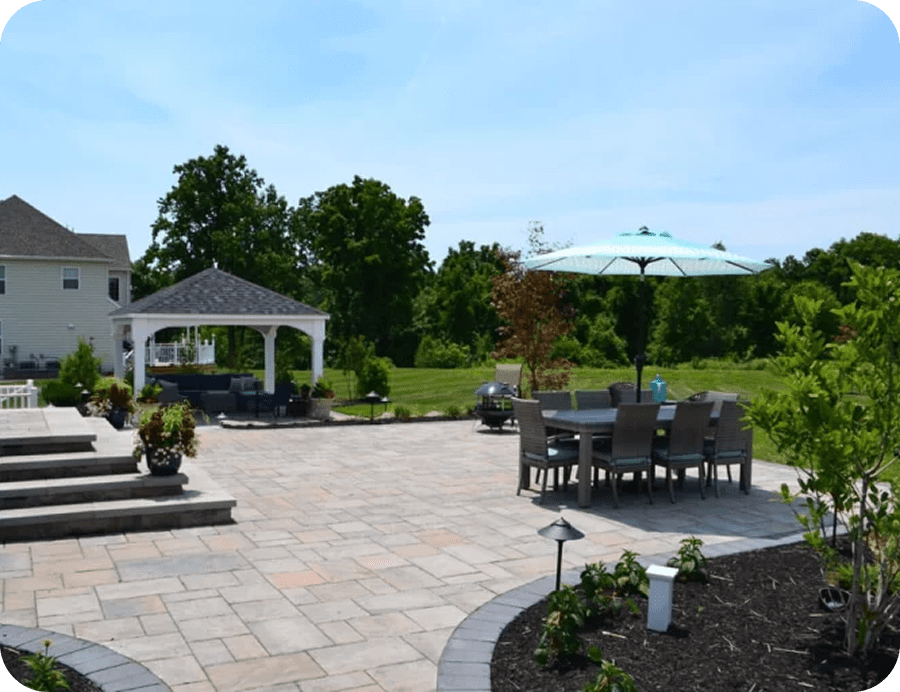 A spacious backyard patio with stone pavers, outdoor dining table and chairs under a large umbrella, a white gazebo, landscaped plants, and green trees in the background.