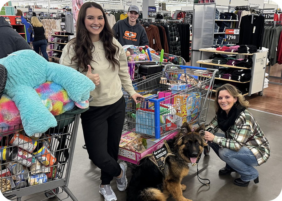 Three people smile in a store next to full shopping carts, with one person giving a thumbs up and another kneeling beside a German Shepherd dog on a leash. Shelves of clothes and toys are visible in the background.