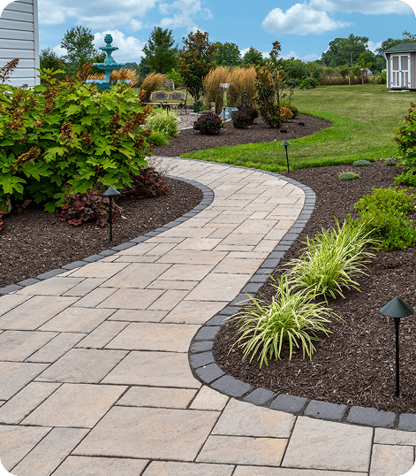 A curved stone walkway bordered by dark bricks winds through landscaped gardens with green shrubs, ornamental grasses, mulch, and solar path lights, leading to a patio area in a spacious yard.