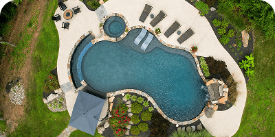 Aerial view of a uniquely shaped backyard swimming pool with stone borders, attached hot tub, sun loungers, patio chairs, umbrella, and surrounding landscaping on a grassy area.