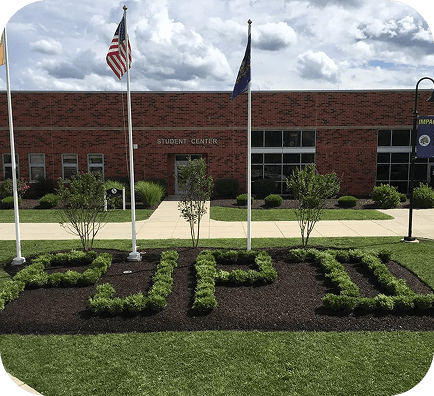 A manicured garden in front of a brick building spells out IMPACT with greenery; three flagpoles stand nearby, and a sign reads STUDENT CENTER. The sky is partly cloudy.