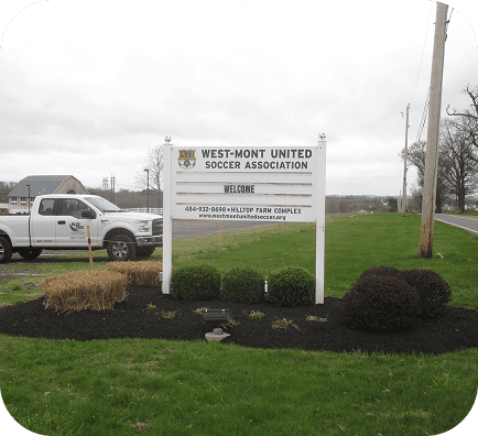 A white sign for the West-Mont United Soccer Association stands on a grassy area by a road, with a white pickup truck and houses in the background. The sign welcomes visitors and provides contact information.