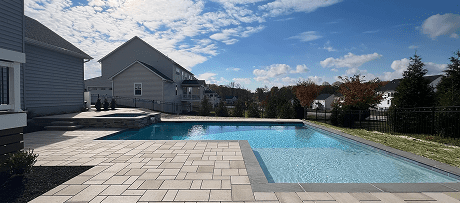 A modern backyard pool with clear water, surrounded by light-colored stone tiles, sits next to a house under a partly cloudy sky in a suburban neighborhood. Trees and houses are visible in the background.