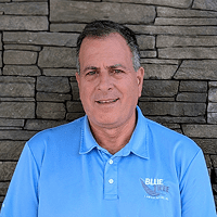 A man in a light blue Blue Title polo shirt stands in front of a stone wall, smiling slightly at the camera.