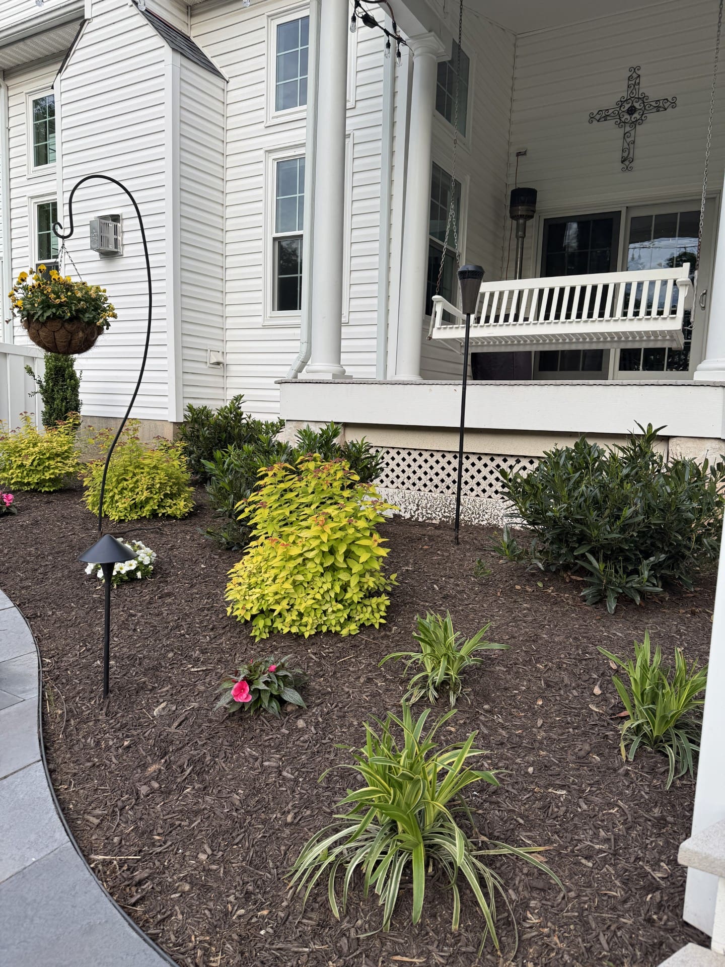 Freshly mulched garden bed with green shrubs and a flowering plant in front of a white house with tall windows, columns, a porch swing, hanging planters, and decorative cross on the wall.