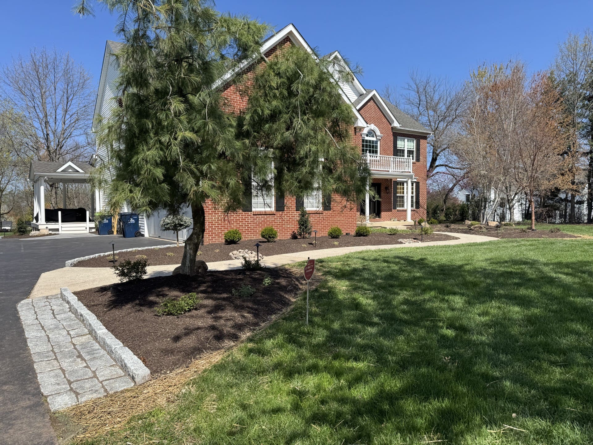 A two-story brick house with white trim, surrounded by neatly landscaped gardens, green grass, and several trees, under a clear blue sky on a sunny day.