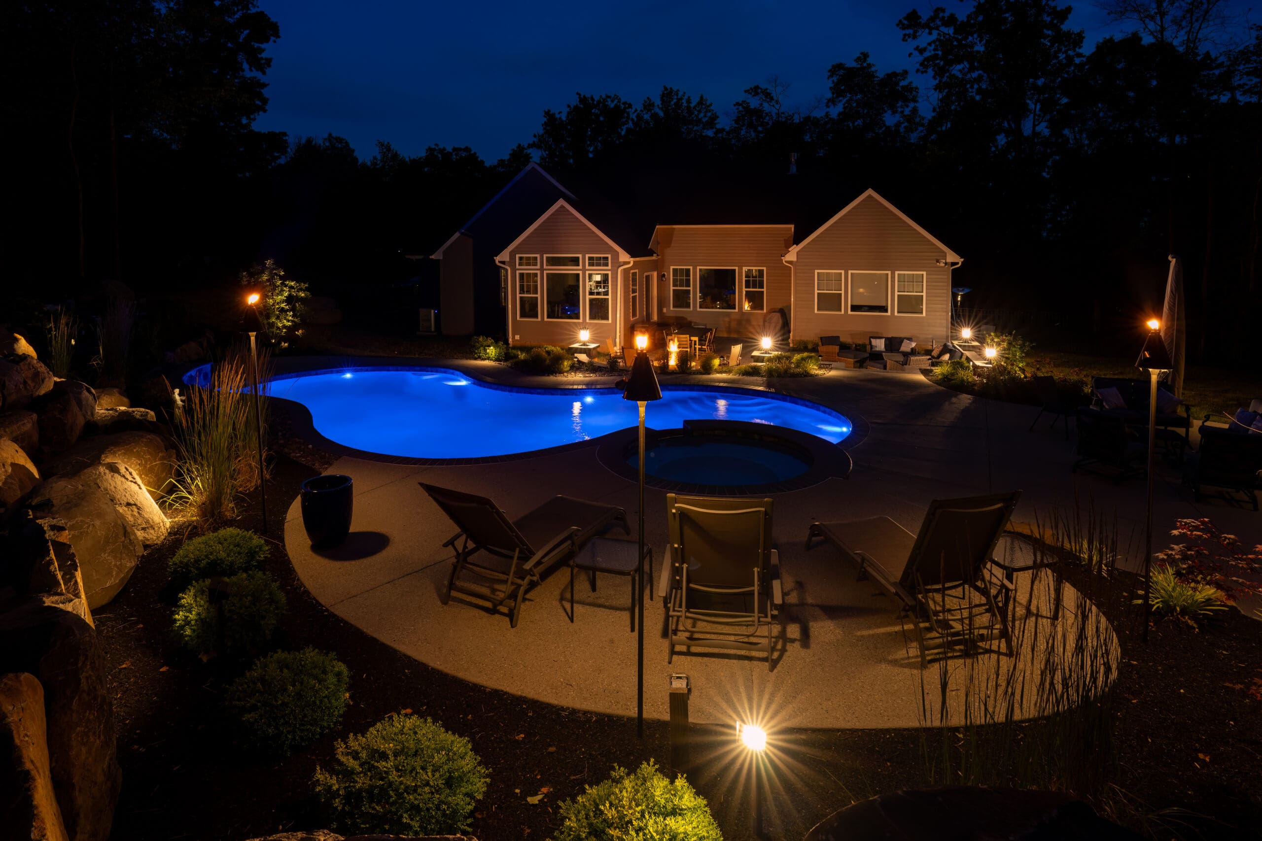 A backyard at night featuring a glowing, illuminated pool and hot tub surrounded by lounge chairs, landscaping, and tiki torches, with a well-lit house in the background.