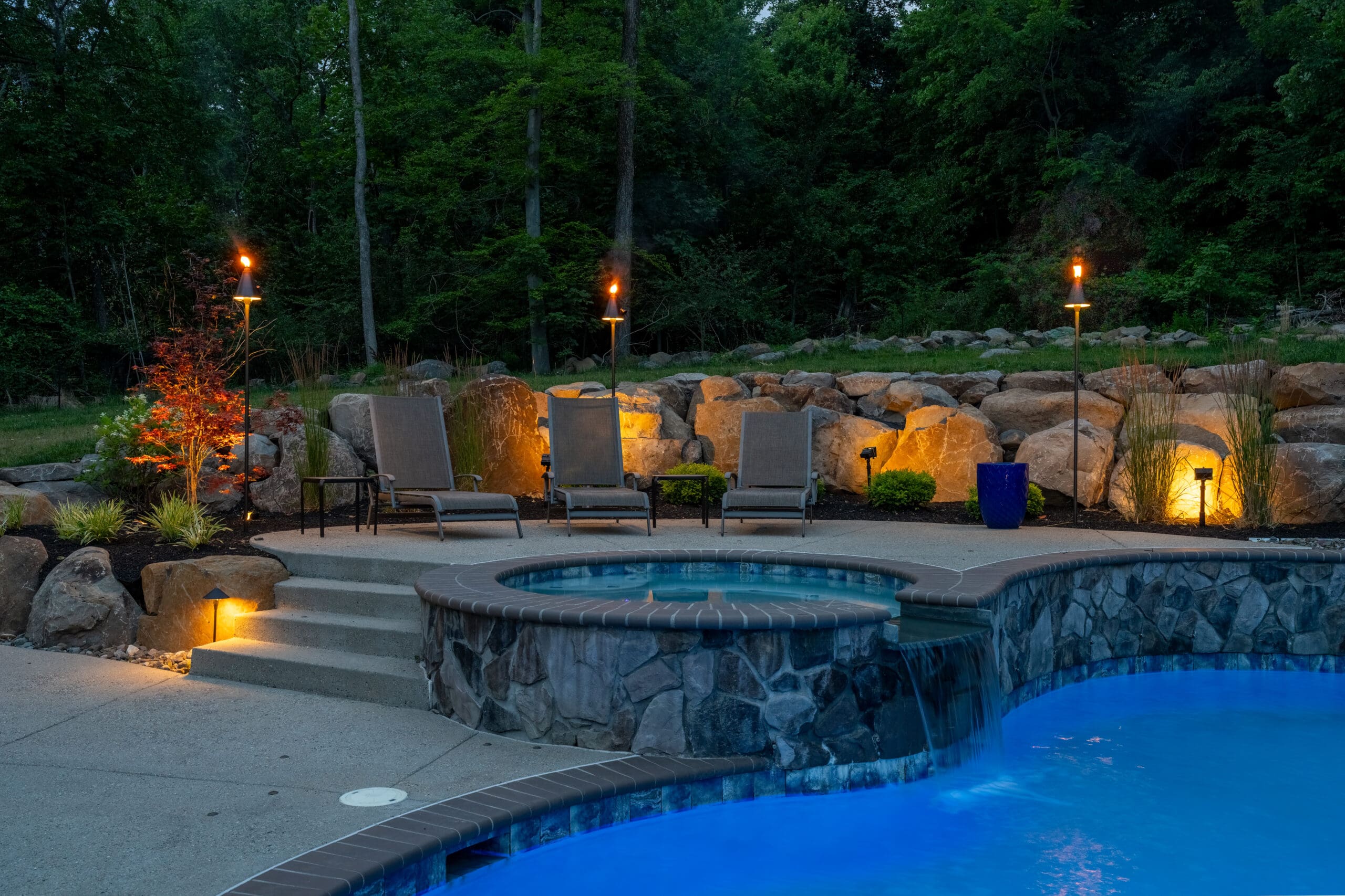A backyard pool and hot tub are lit at dusk, surrounded by lounge chairs, glowing tiki torches, and landscape lighting with trees and rocks in the background, creating a relaxing atmosphere.