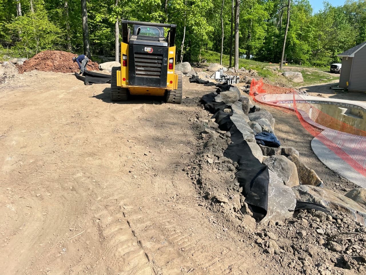 A yellow skid steer loader is parked on a dirt construction site near a curved line of large rocks and black fabric. A person works by the rocks, and trees and a house are visible in the background.