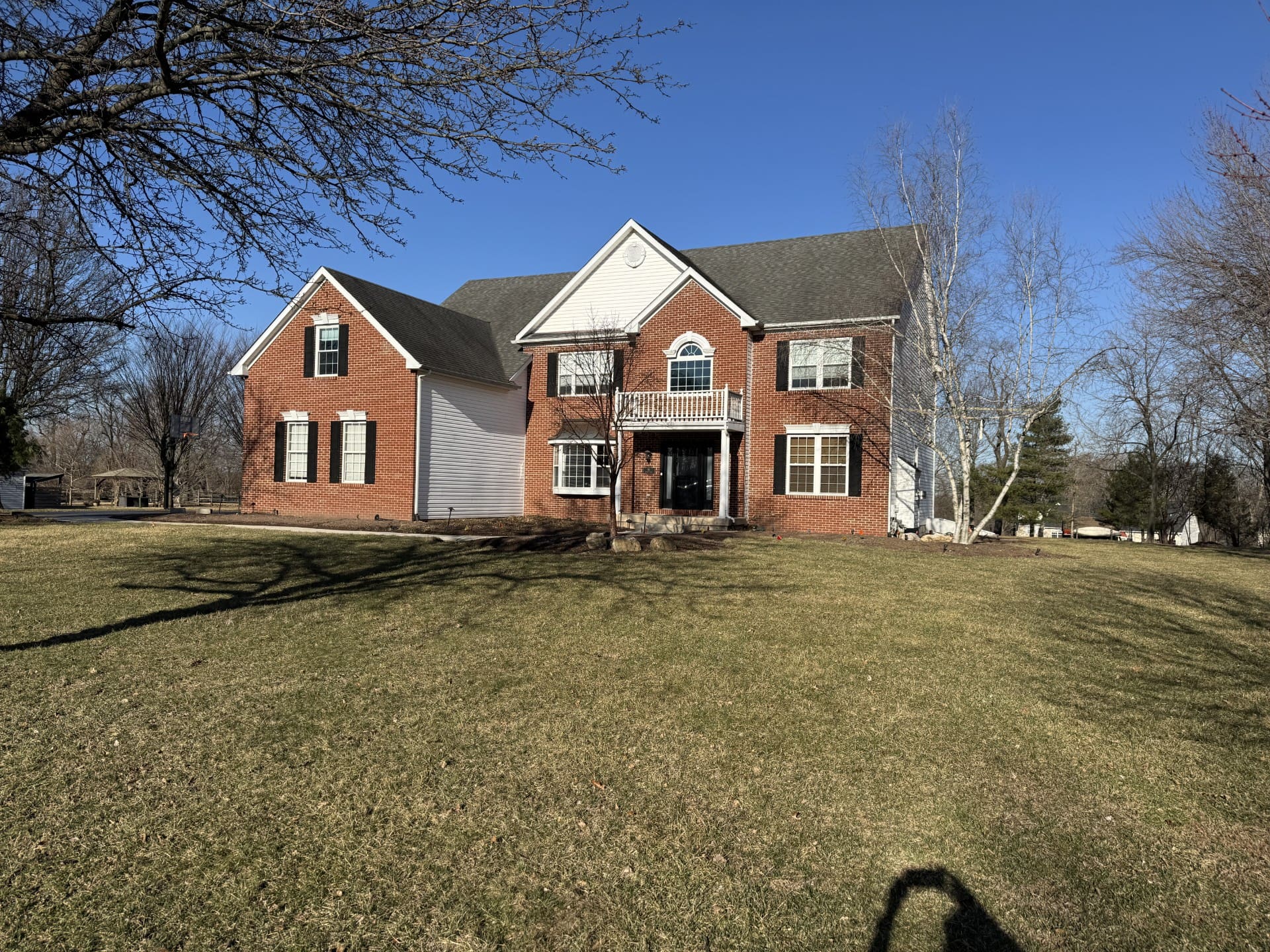 A large two-story brick and white-sided house with multiple windows sits on a spacious lawn under a clear blue sky, with bare trees and shadows visible in the foreground.