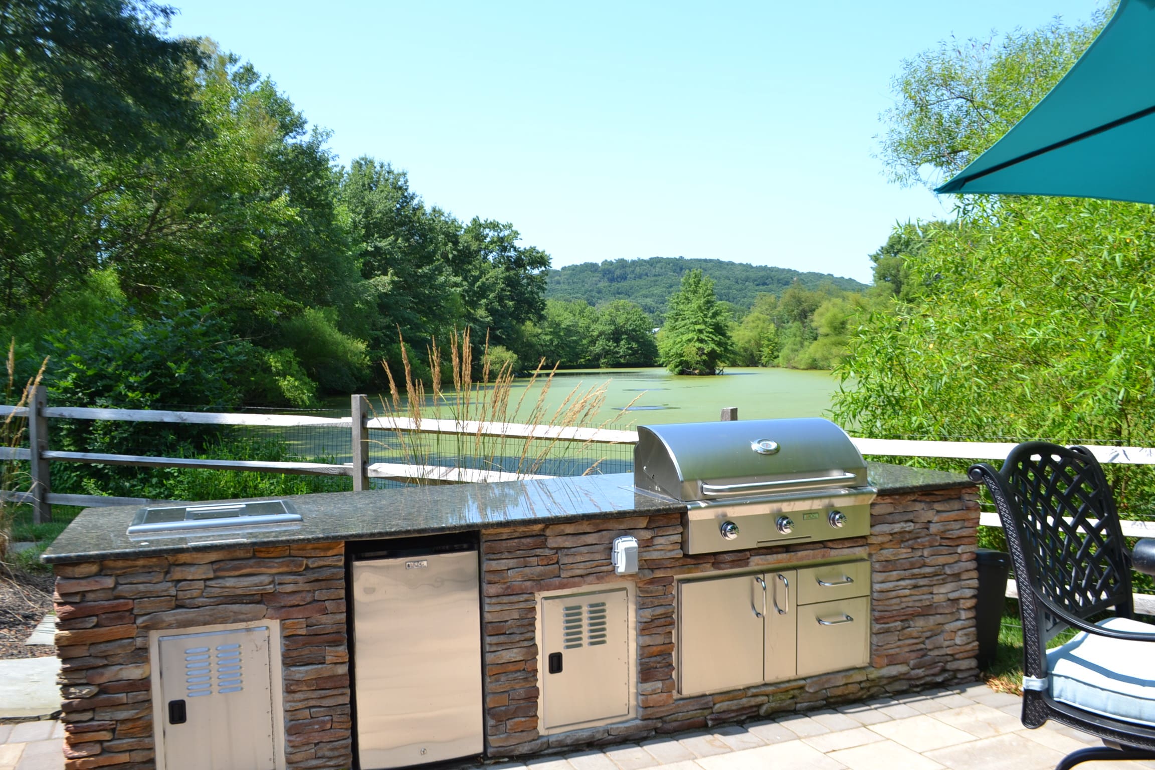 Outdoor kitchen with a stone countertop, stainless steel grill, and appliances, set on a patio overlooking a pond, green trees, and hills under a clear blue sky. A fence and patio furniture are also visible.