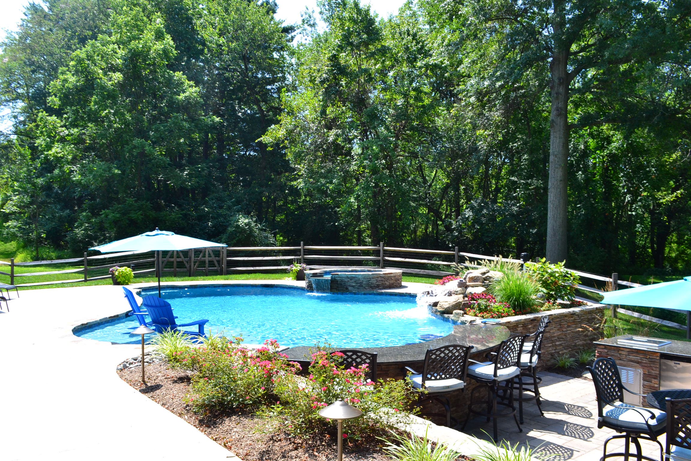 A backyard pool with waterfall features, surrounded by trees and a wooden fence. Patio chairs and tables with umbrellas are arranged nearby, and landscaped plants border the pool area.