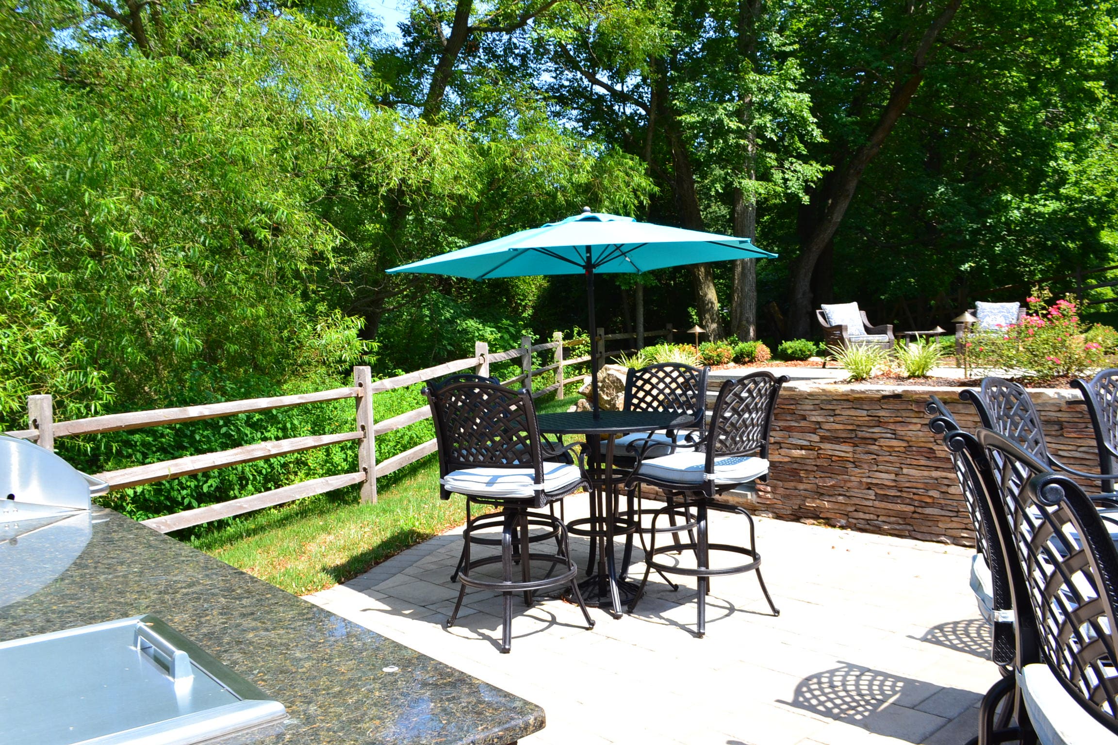 A sunny outdoor patio features a table with four chairs under a blue umbrella, stone retaining wall, lush green trees, and a wooden fence in the background.