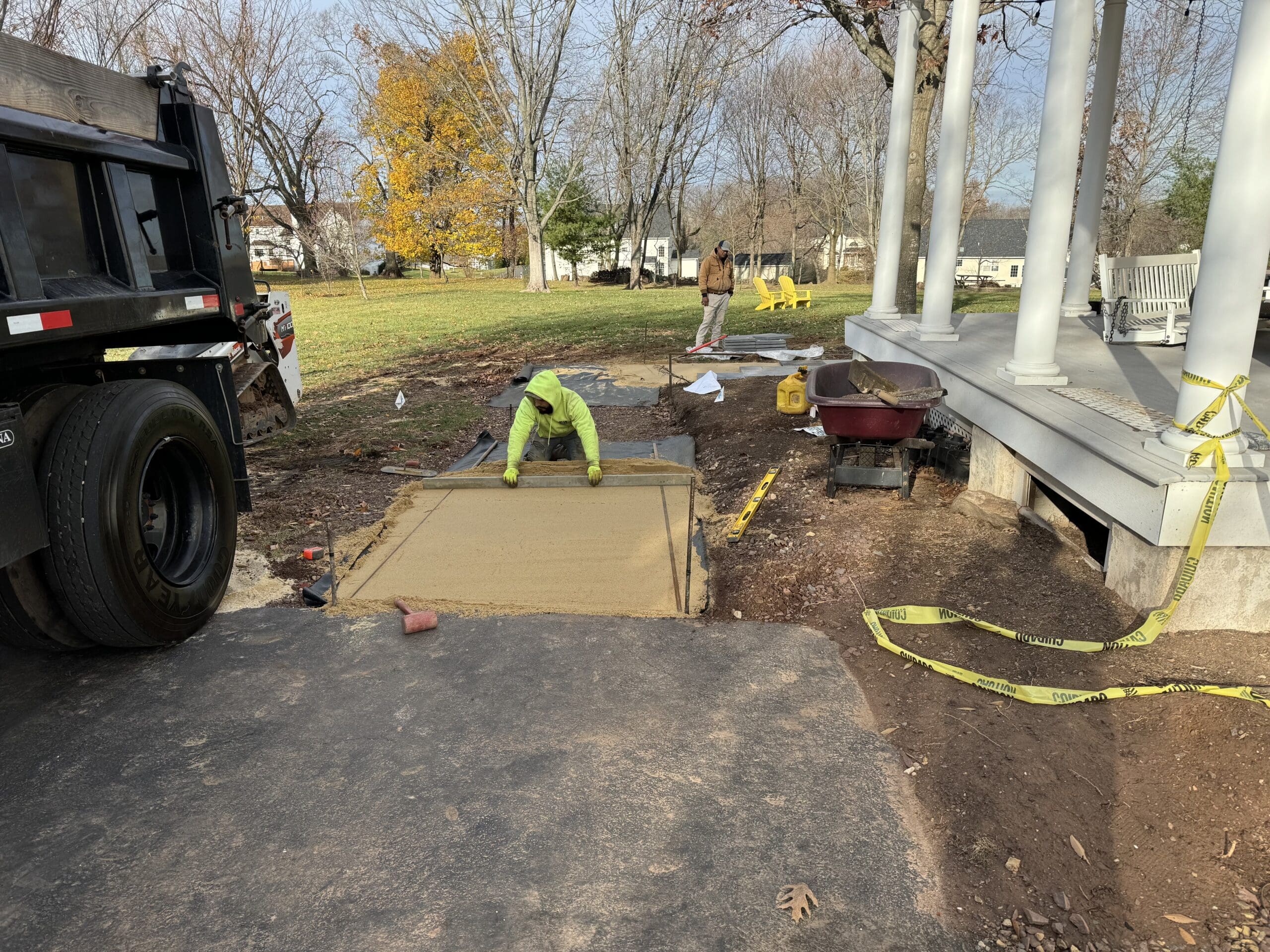 A construction worker spreads sand to prepare a rectangular section of ground for pavement near a building with white columns. Tools, caution tape, and a wheelbarrow are nearby, and trees are in the background.