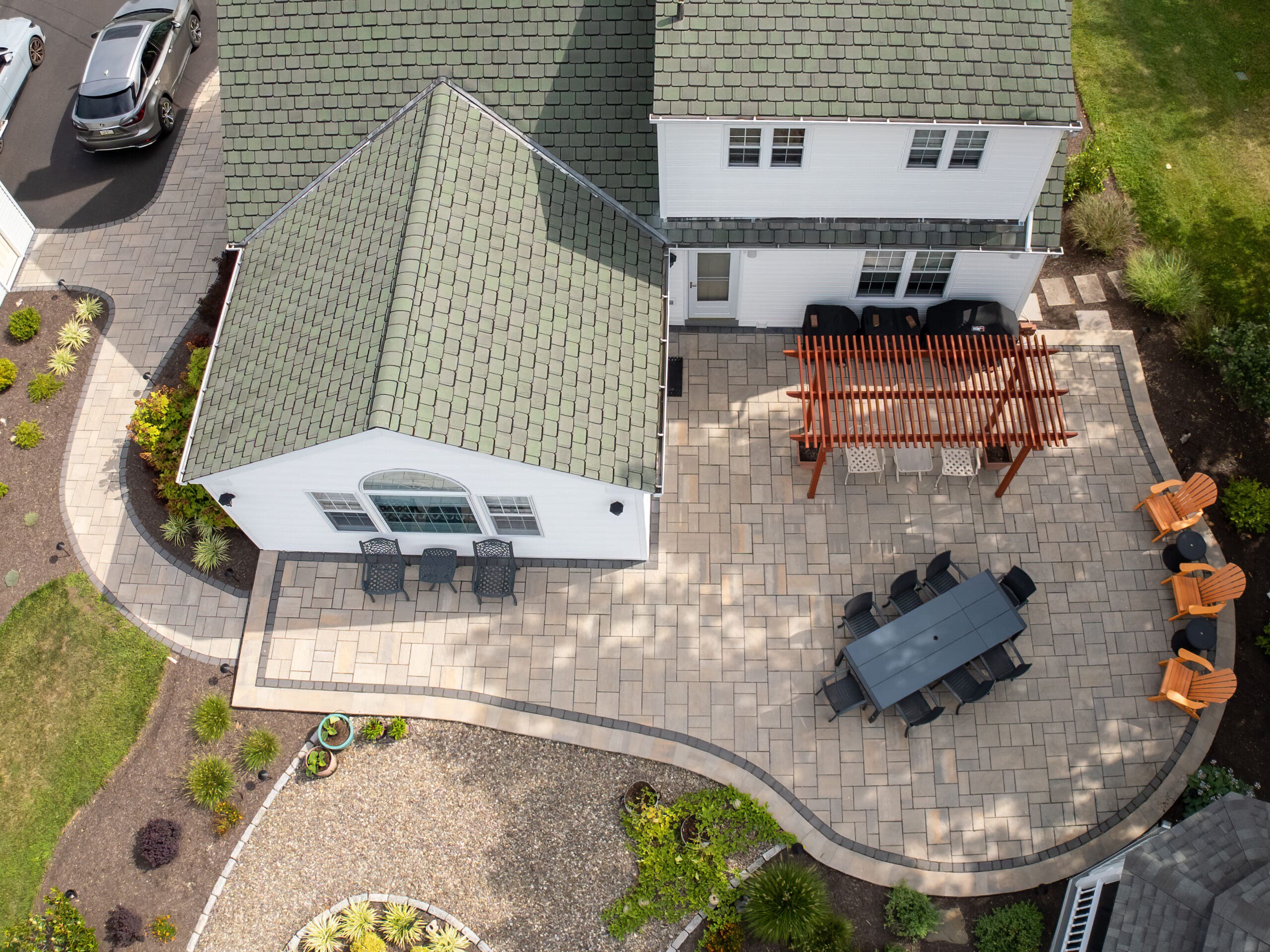 Aerial view of a backyard patio with light-colored pavers, a dining table with chairs, an outdoor pergola, lounge chairs, and landscaped garden beds surrounding the paved area.