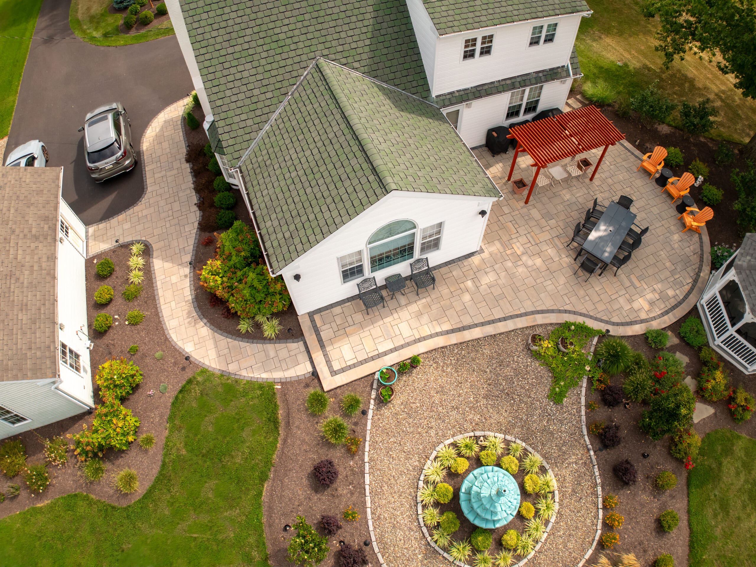 Aerial view of a house with a stone patio, outdoor dining area, orange chairs by a pergola, landscaped garden, and a circular gravel area with a decorative fountain. Two cars are parked nearby.
