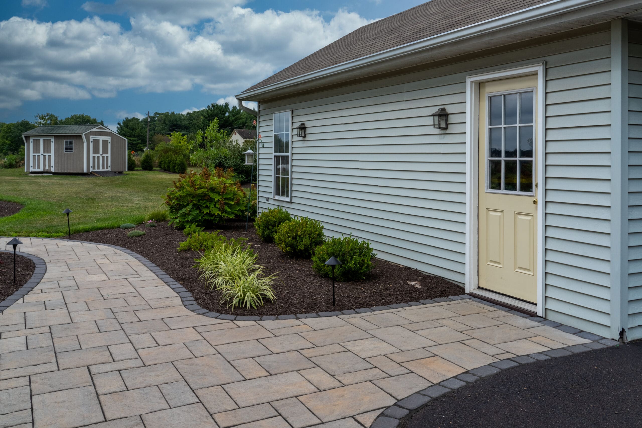 A paved walkway curves past landscaped bushes and leads to a beige door on a light gray house, with a small shed and green lawn visible in the background under a partly cloudy sky.