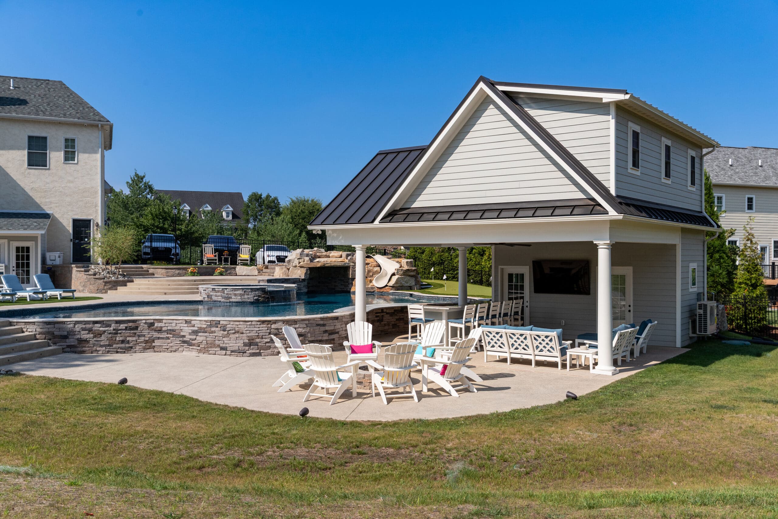 A backyard with a swimming pool, hot tub, and slide next to a covered patio area. White Adirondack chairs and tables are arranged on the patio, and there’s a two-story pool house nearby. The sky is clear and blue.