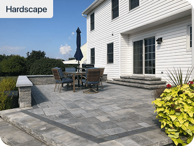 Patio area with gray stone pavers, outdoor table, chairs, and umbrella next to a white house. Steps lead to glass doors, with green plants and flowers along the edge, labeled Hardscape in the corner.