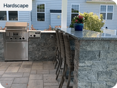 Outdoor kitchen with a built-in stainless steel grill and bar area with wicker barstools, stone countertops, and a potted plant on the counter; house and greenery in the background.
