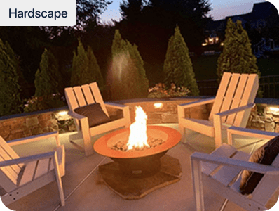Four white wooden chairs surround a stone fire pit with a bright flame on a patio, bordered by stone walls and tall evergreen trees, during dusk.