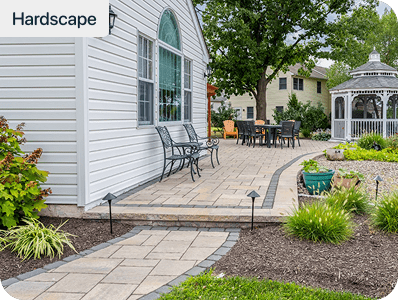 A paved stone walkway and patio area beside a white house, featuring benches, outdoor dining furniture, landscaping with shrubs, and a white gazebo in the background.