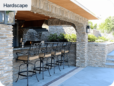 Outdoor stone patio with a built-in bar counter, four ornate metal barstools, stone pillars, and landscaped greenery in the background. Hardscape is written in the top left corner.