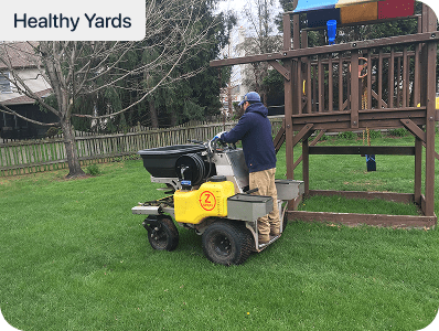 A person operates a yellow lawn care machine on grass near a wooden playset. Trees and a fence are visible in the background. The image is labeled Healthy Yards in the top left corner.