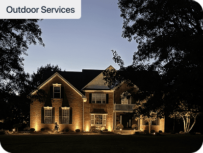 A large, two-story brick house with exterior lights illuminating the front at dusk. Trees frame the yard, and the sky is darkening. A label in the top left corner reads Outdoor Services.