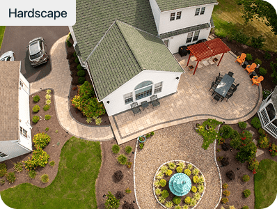 Aerial view of a homes backyard with stone walkways, patio seating, a pergola, green lawn, landscaped garden beds, and a circular fountain centerpiece. Hardscape text appears in the top left corner.