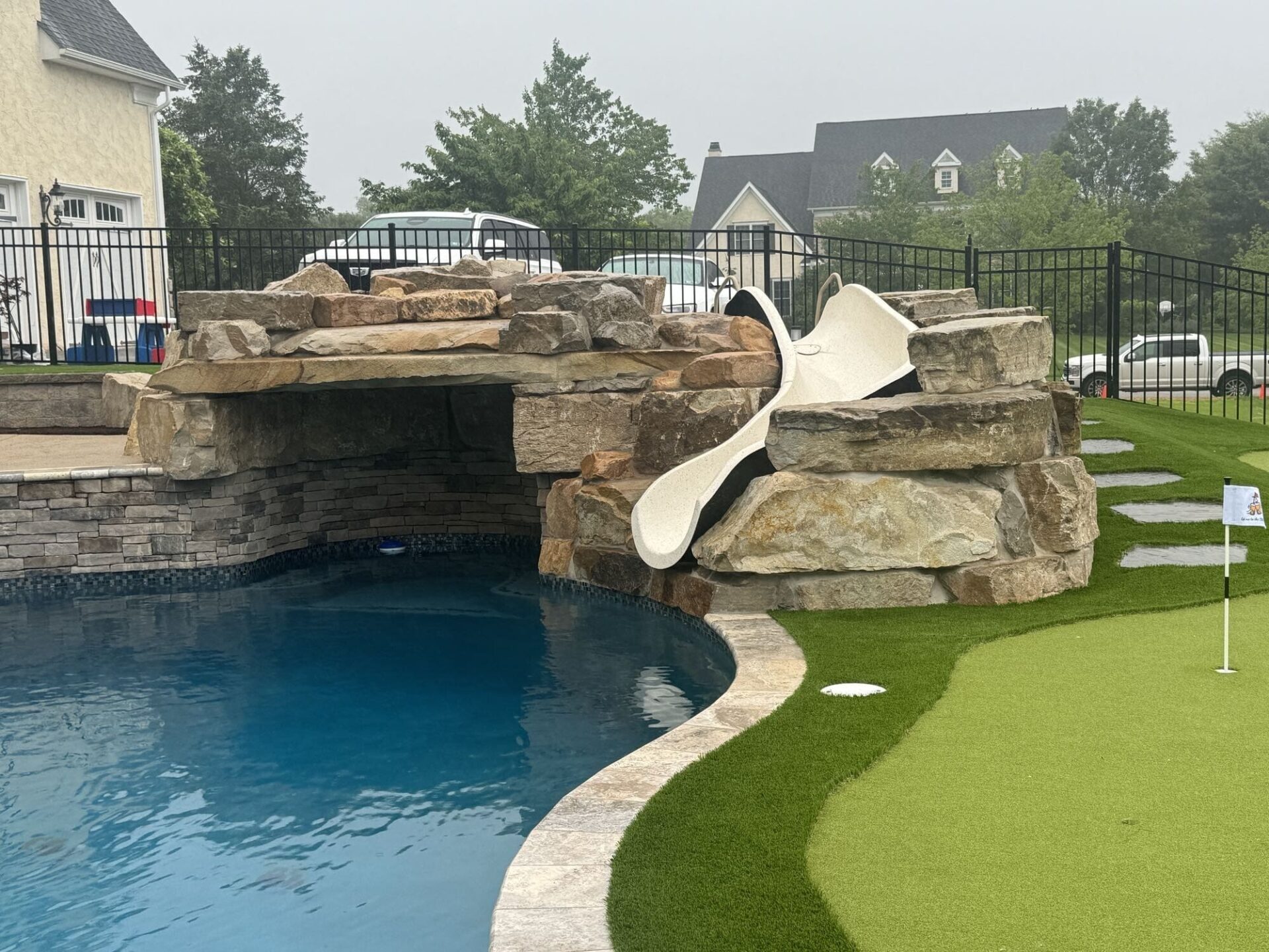 A backyard scene with a swimming pool featuring a rock waterfall, a white slide, and an artificial turf putting green with a small flag, surrounded by a black metal fence and houses in the background.