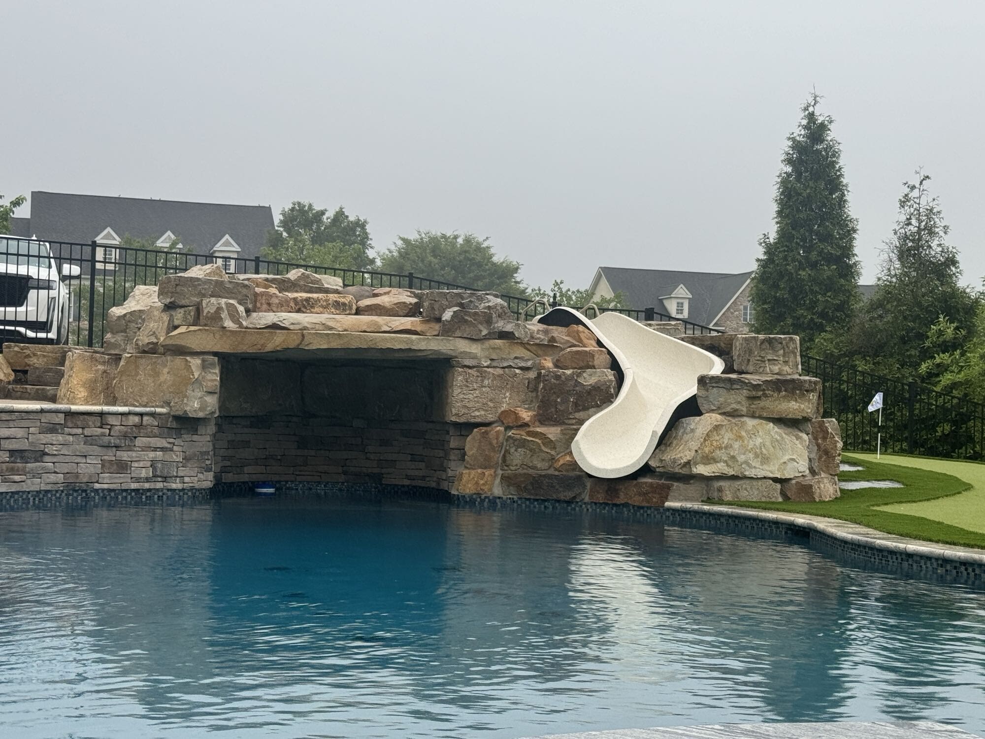 A backyard pool with a rock grotto and a white slide curving into the water. There is a small putting green with a flag in the background, and houses are visible beyond the pool area.