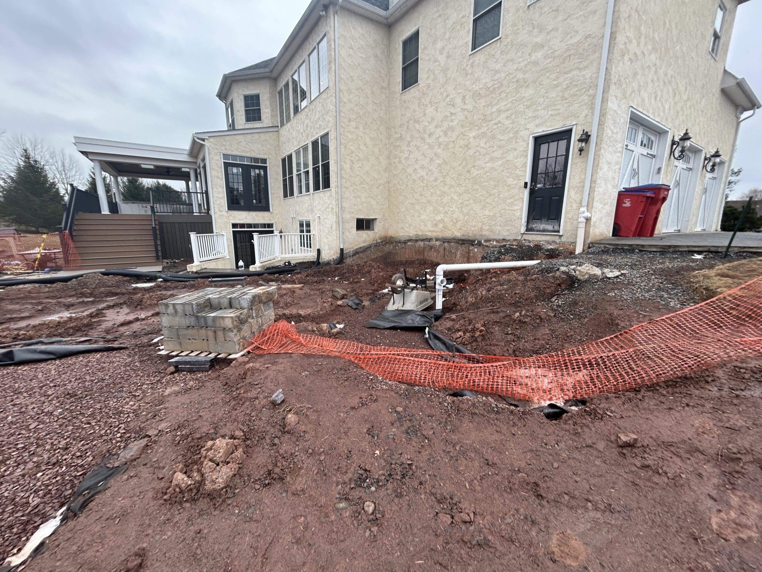 A house with a beige exterior stands next to a construction site. Exposed dirt, stacked concrete blocks, pipes, and an orange safety fence are visible, indicating ongoing landscaping or renovation work.