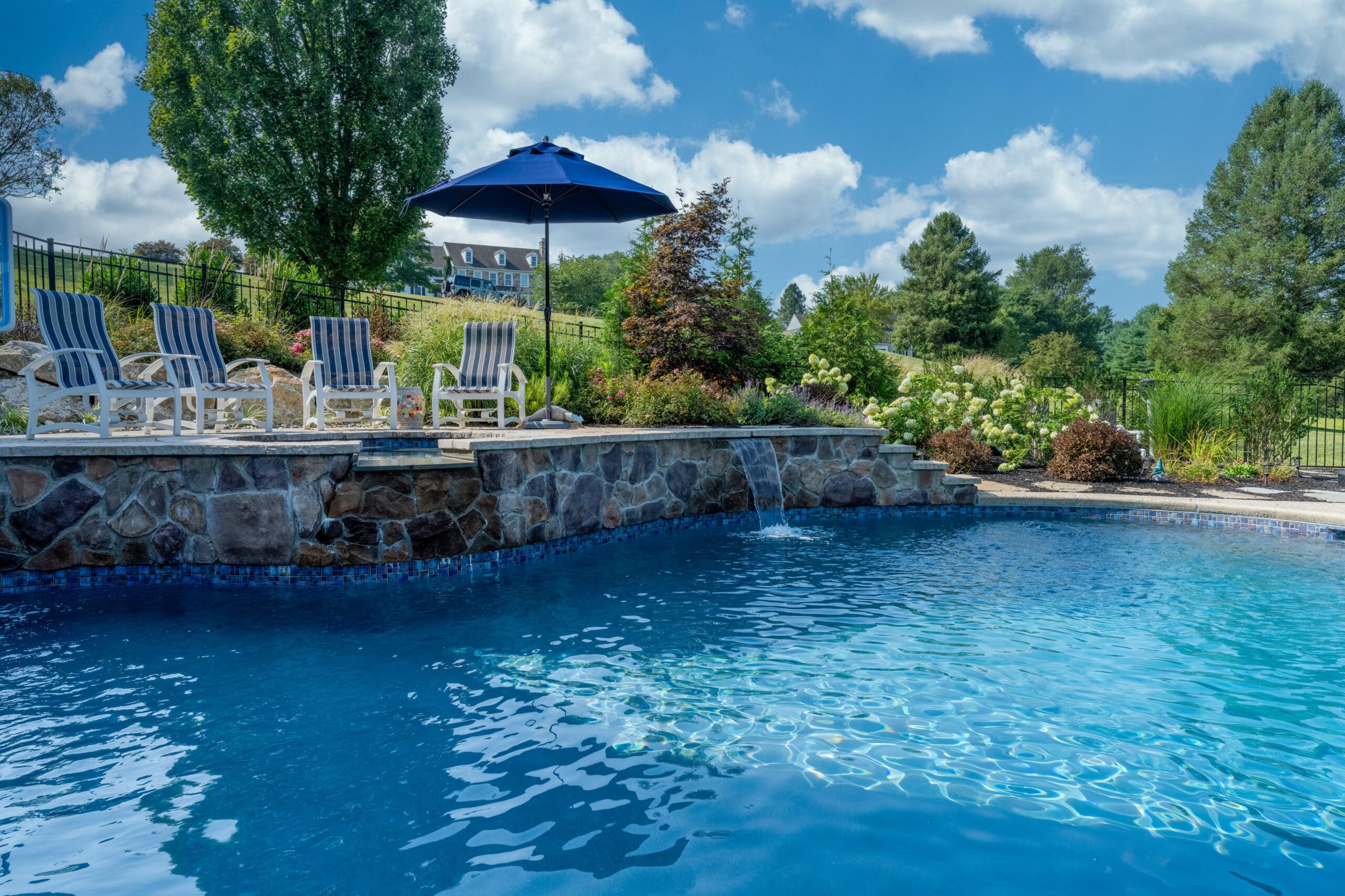 A sparkling blue swimming pool with water features, surrounded by stone, lush landscaping, lounge chairs, and a navy umbrella under a sunny sky with scattered clouds and tall trees in the background.