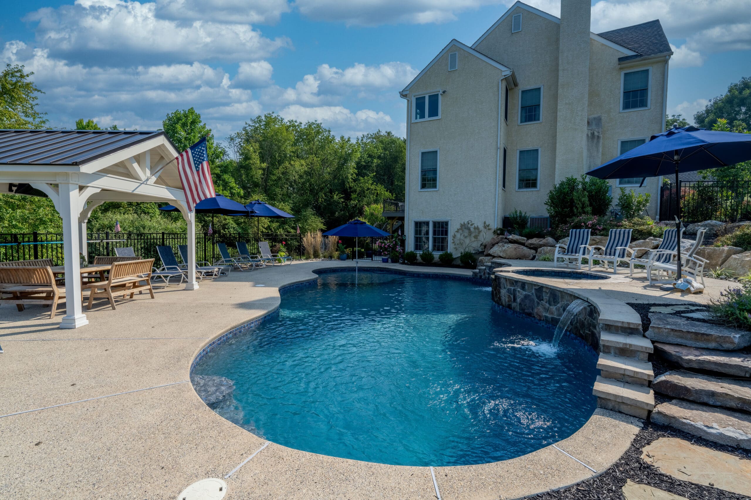 Backyard swimming pool with a small waterfall feature, surrounded by lounge chairs and blue umbrellas, next to a two-story house. A covered patio area with seating and an American flag are also visible.