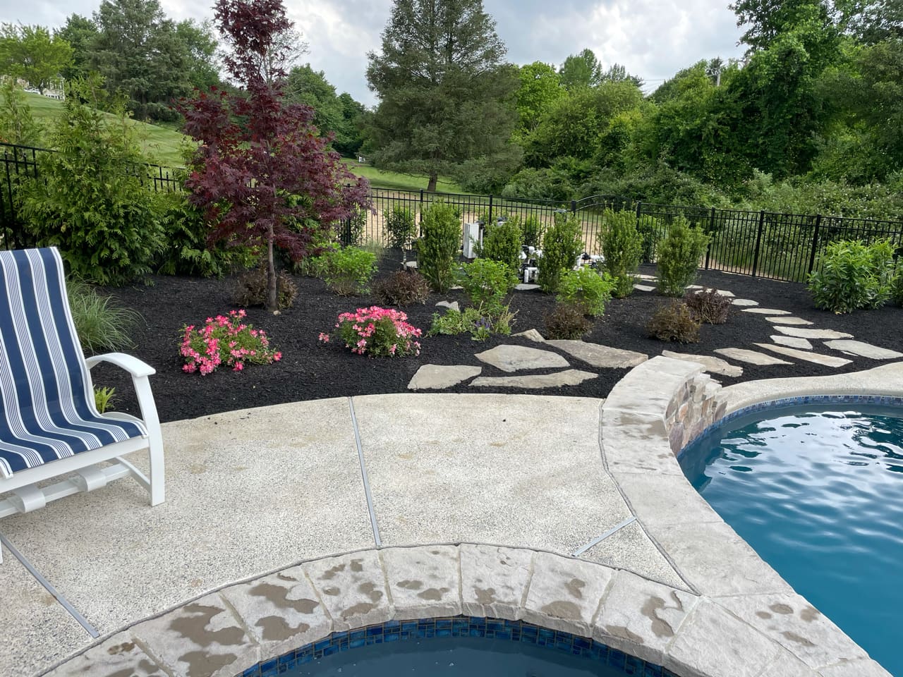 A backyard poolside area with a curved stone patio, striped lounge chair, landscaped flower beds, mulched garden, stone path, and trees with a black metal fence in the background.