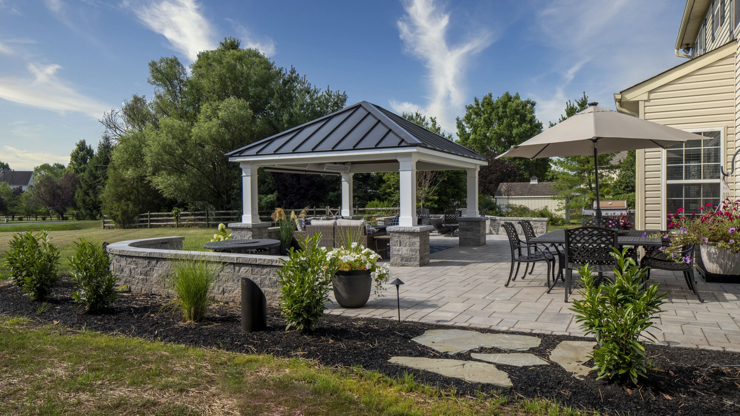 A backyard patio with a covered seating area, outdoor dining table with umbrella, potted plants, stonework, and landscaped greenery under a blue sky with scattered clouds.