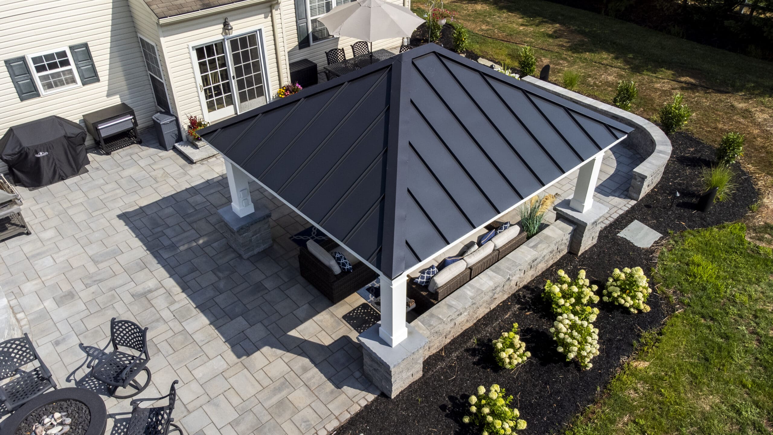 A backyard patio with a modern black metal-roofed pavilion, outdoor furniture, pavers, and landscaped garden beds with shrubs and flowers, adjacent to a light-colored house.