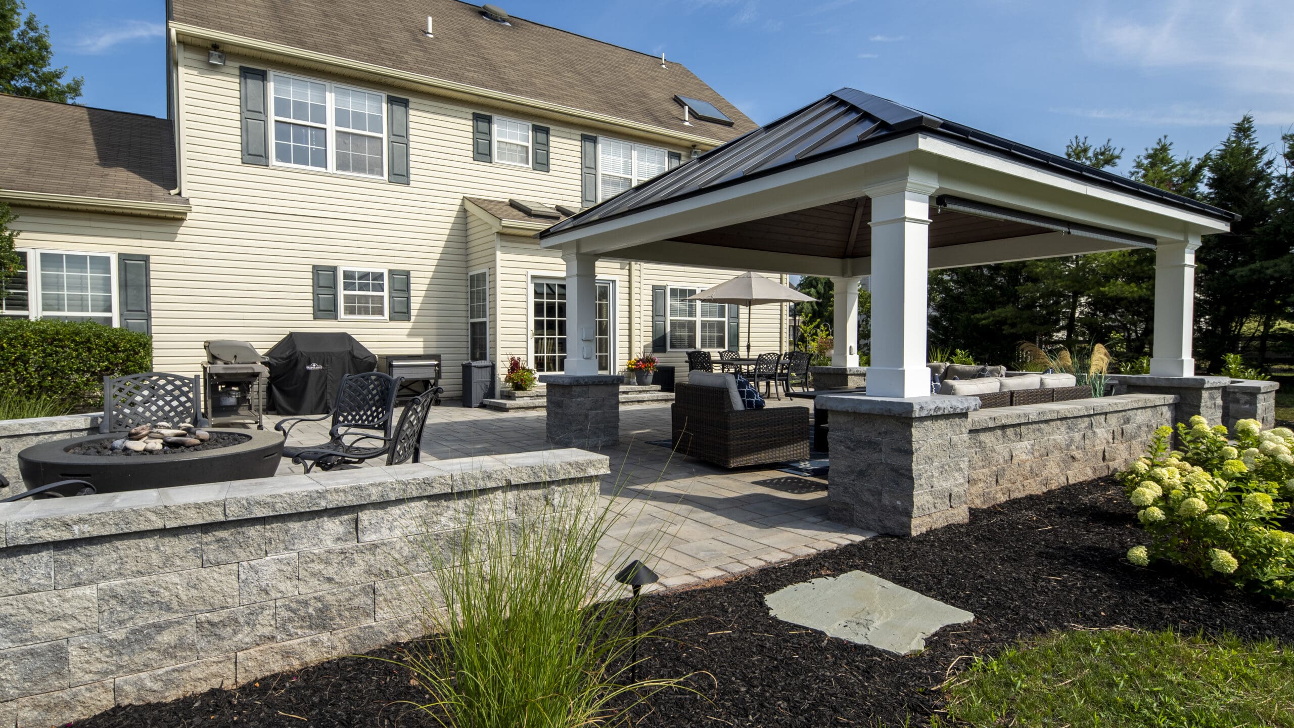 Spacious backyard patio with a stone fire pit, black metal chairs, a covered seating area, grill, and lush landscaping beside a beige two-story house under a blue sky.