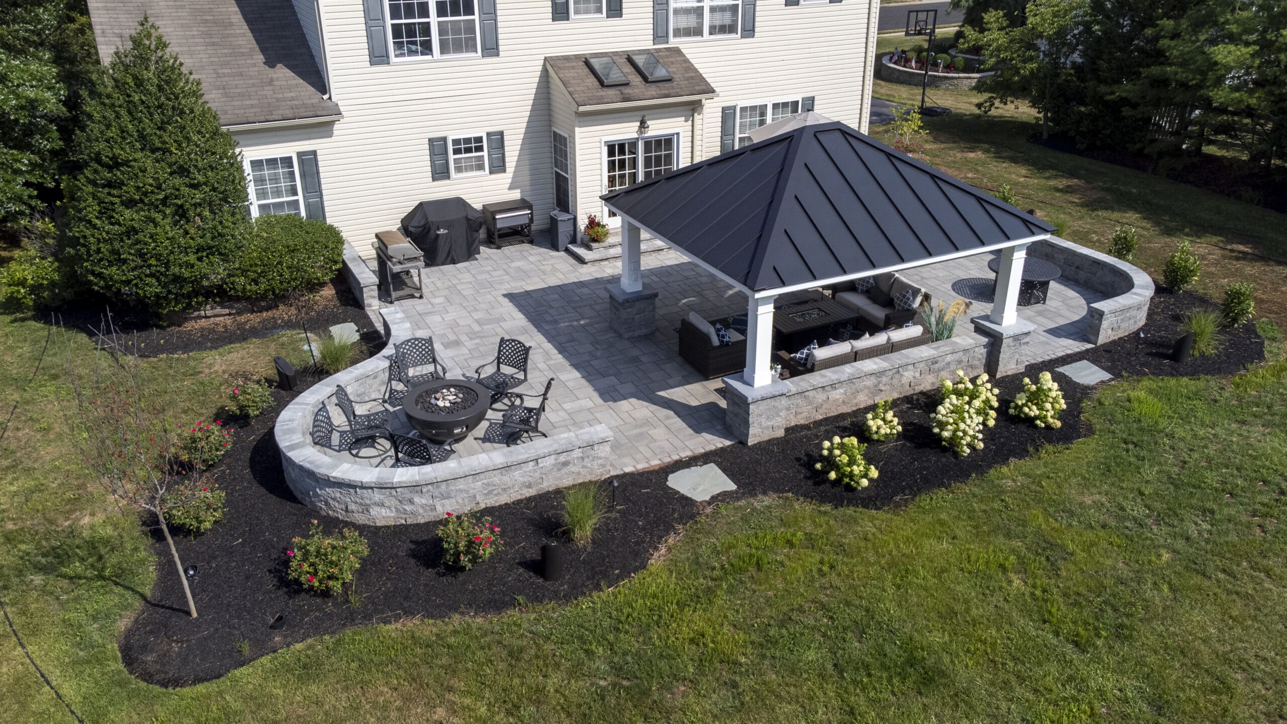Aerial view of a spacious backyard patio featuring a covered seating area, outdoor dining set, grill, fire pit, and surrounding landscaping with shrubs, flowers, and small trees.