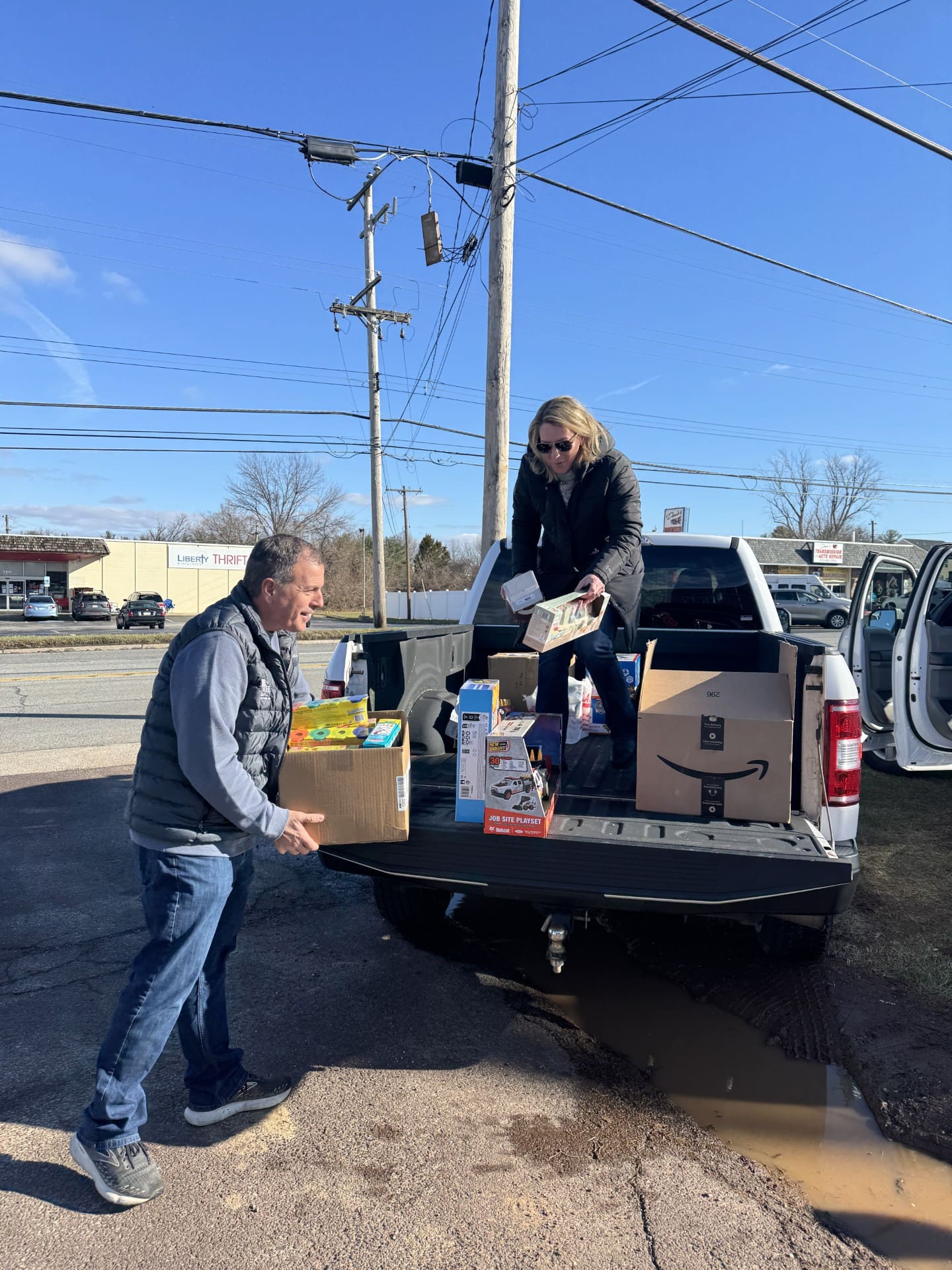 Two people unload boxes of groceries and household items from the bed of a pickup truck on a sunny day. One person stands on the ground holding a box; the other stands in the truck bed handing down items.